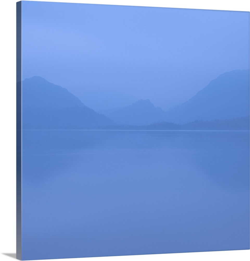 Square photograph of a blue foggy lake with mountains in the background.