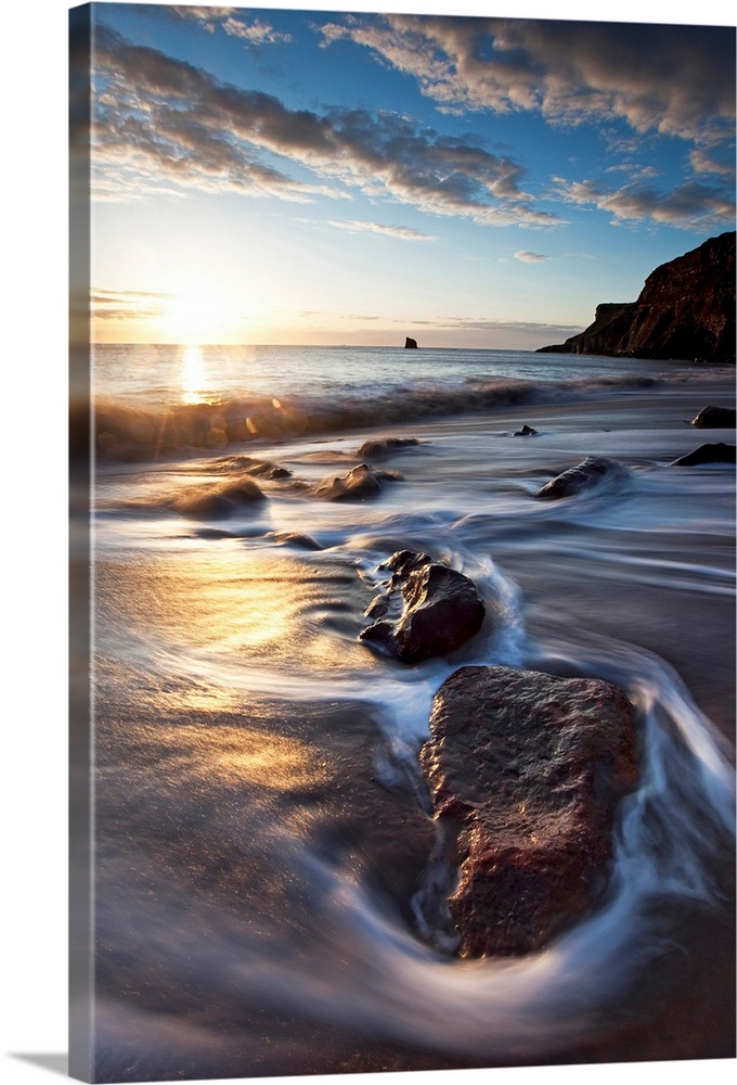 Vertical photograph on a large canvas of the sun rising over waters rushing onto a rocky beach, in Tyneside, Northern Engl...