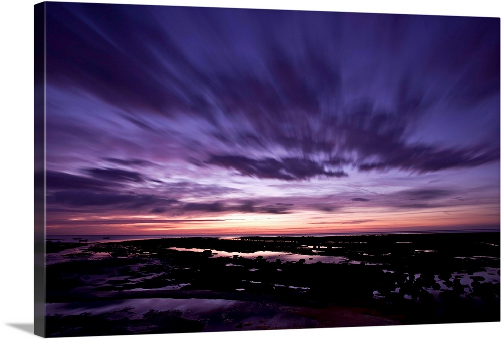 A dramatic seascape at dawn with deep purple magenta pink skies and clouds over wet sands.
