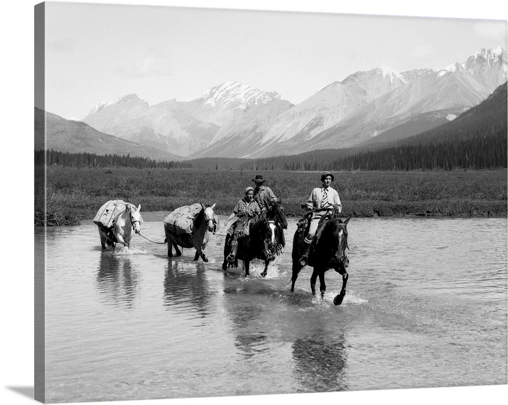 1930s Two Women Man Riding Horseback Through Shallow Water Two Extra Horses Carry Camping Equipment Mountains In Background.