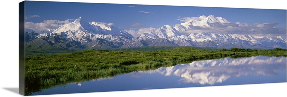 A snow covered mountain range is photographed in panoramic view with a large field shown just in front and a body of water...