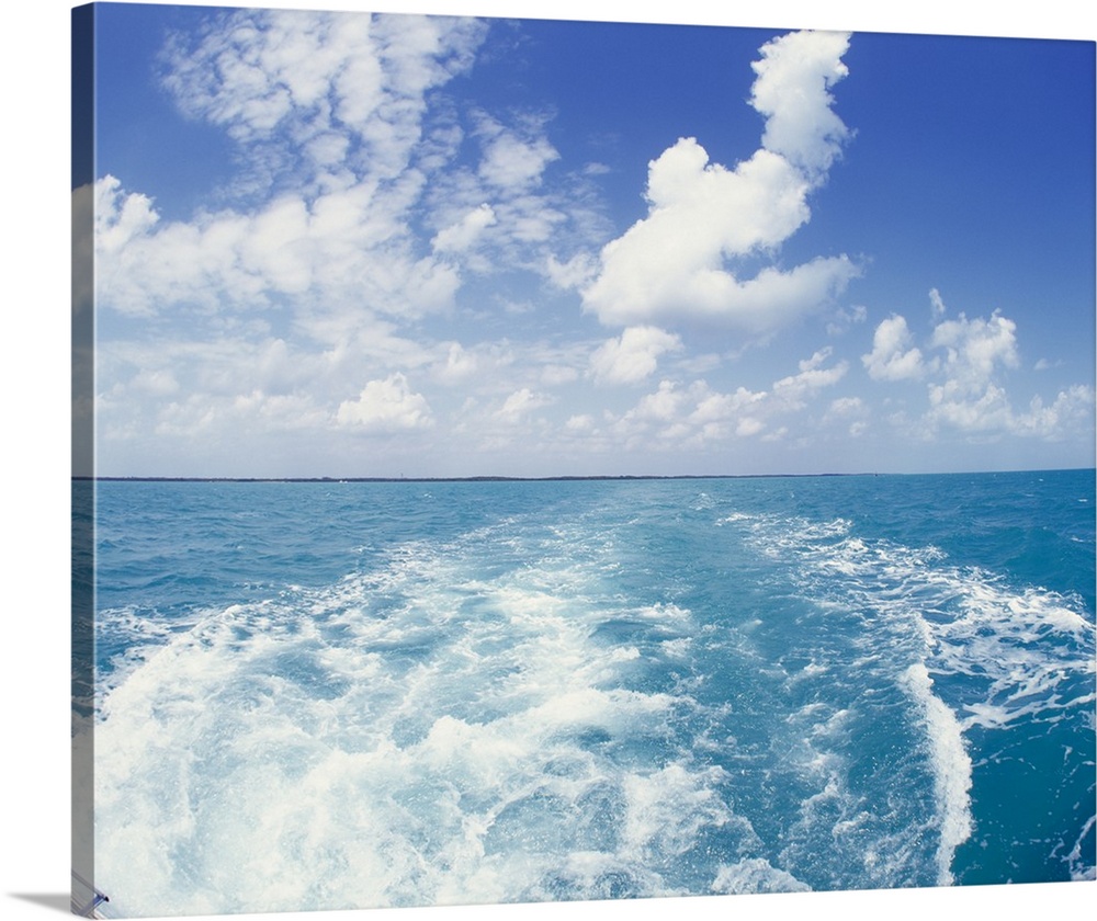 Photo on canvas of the ocean with land in the far distance and puffy clouds above looking from behind a running boat.