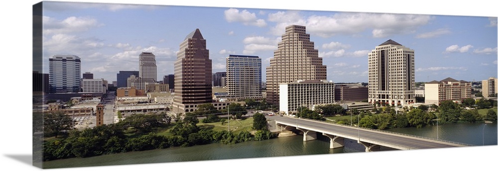 Panoramic photograph of the Austin, Texas skyline beneath a blue sky with small fluffy clouds, the edge of Town Lake in th...
