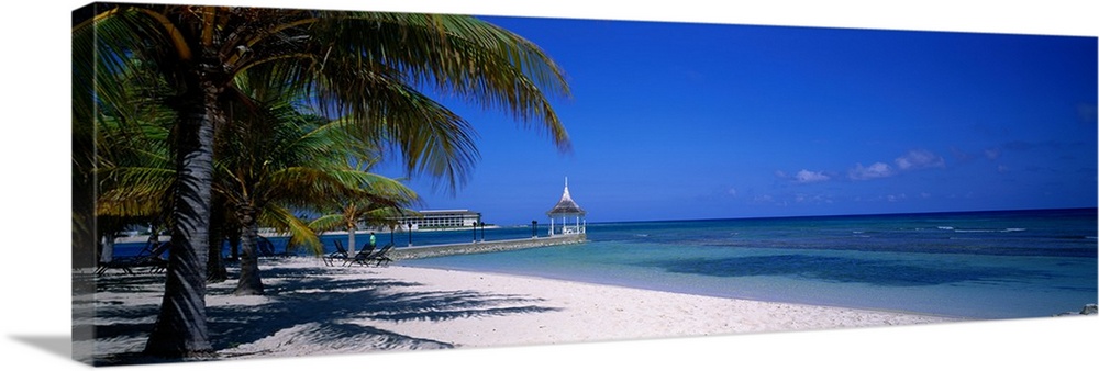 A pier stretches into the tropical ocean next to a white sandy beach and palm trees in the Caribbean.