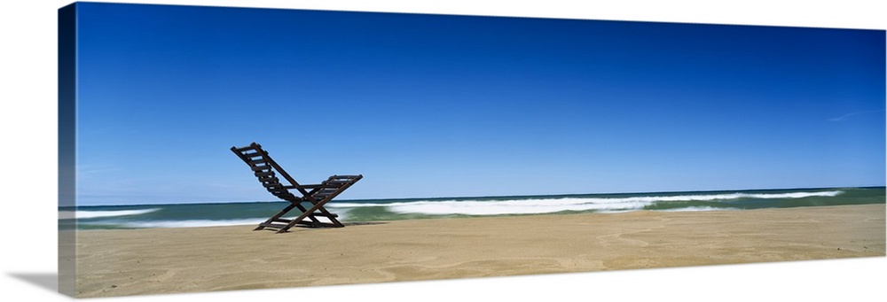 Huge panoramic piece of a lone beach chair skewed to the left sitting in the sand with waves crashing and a clear blue sky...