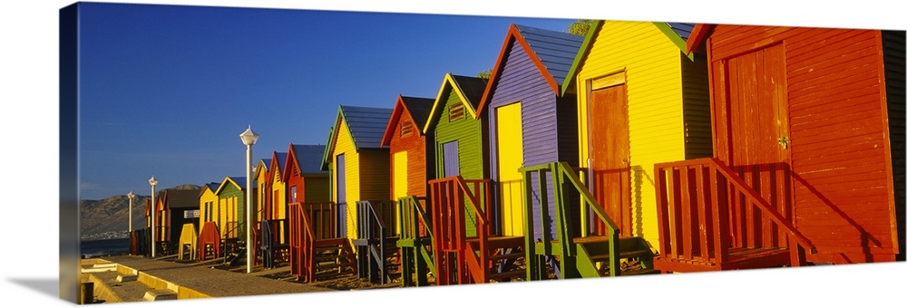 Panoramic photograph displays a line of vibrantly colored seaside shacks on a sunny day.  In the background there is a mou...