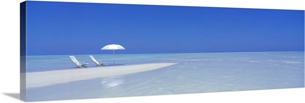 Large panoramic picture of two white chairs and a white beach umbrella sitting on sand that stretches into the crystal cle...