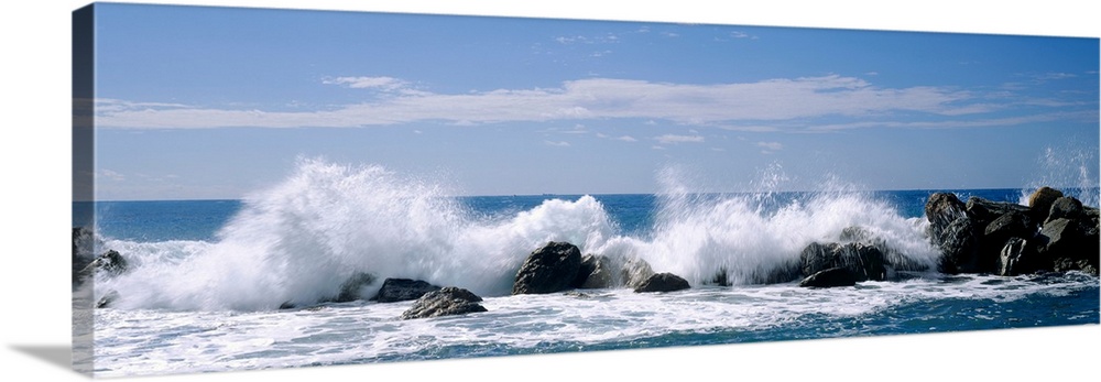 Panoramic photograph of swells crashing against rocks under a cloudy sky.