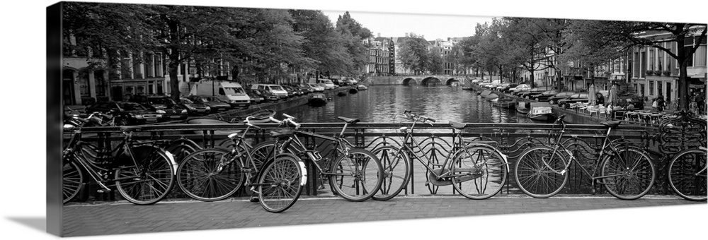 Panoramic monochromatic photograph focuses on a group of bikes resting on an overpass over a river in Europe.  The sides o...