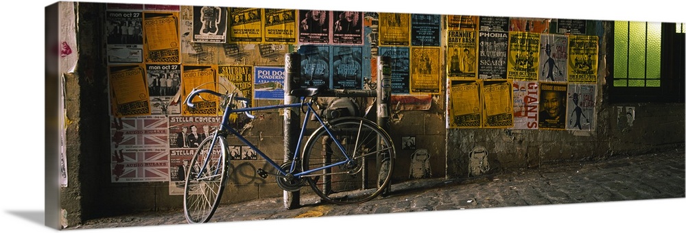 This panoramic photograph is a cobblestoned street build on a slant and a wall covered in band posters with a bike chained...
