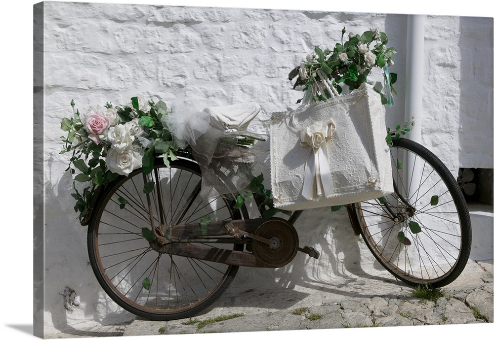 A bike with rusty gears covered in beautiful roses with a decorative ribboned tote bag leaning against a textured wall.
