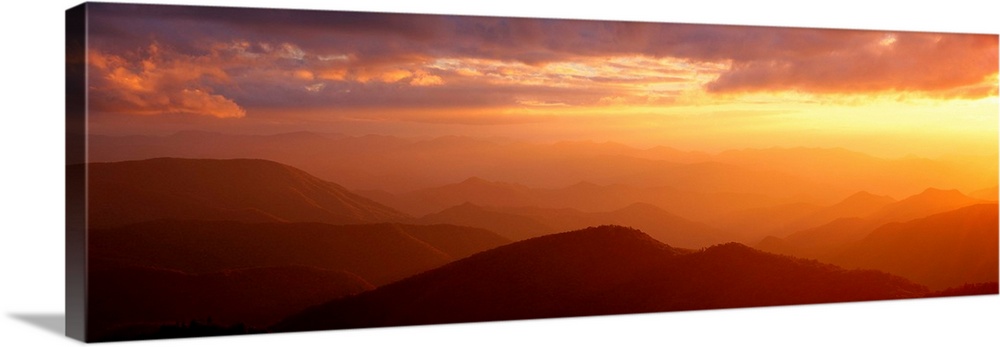 Panoramic photograph shows the glow of the sun as it begins to set on a mountain range within North Carolina.  The warm ra...