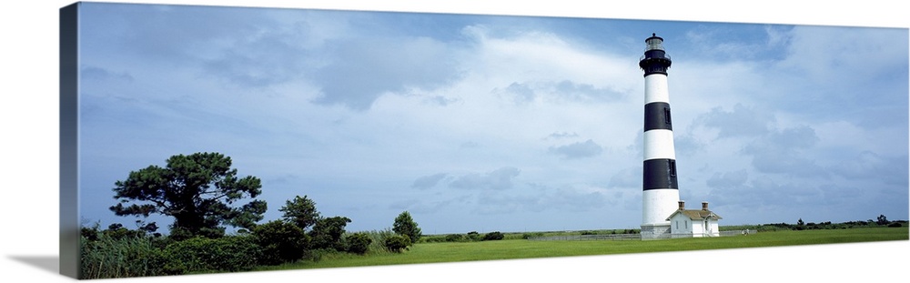 Panoramic photograph of striped Bodie Lighthouse surrounded by a green landscape, beneath a cloudy blue grey sky.