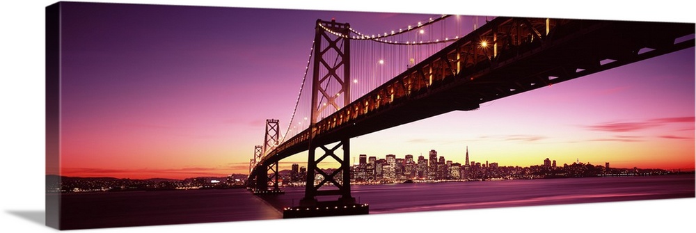 Large panoramic photo print of a long bridge leading to a lit up city off in the distance at sunset.