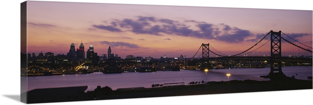 Panoramic photograph of city skyline and overpass at dusk.  The sun is setting, the sky is cloudy, and the buildings are l...