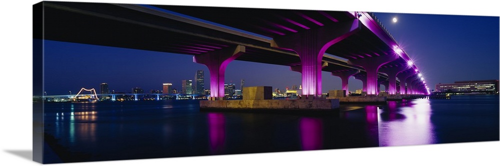 Panoramic photograph taken from the shoreline looking up at the underside of a large bridge illuminating at night and refl...
