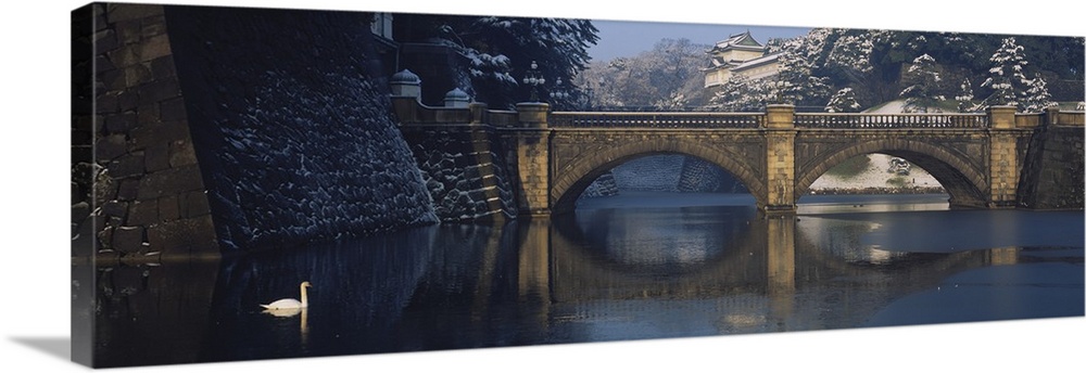 Panoramic photograph of the stone Bridge Nijubashi over calm waters, surrounded by a snow covered landscape as a swan floa...