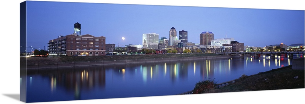 This panoramic photograph is taken of buildings lit up at night that sit on the water front in Des Moines and reflect in t...