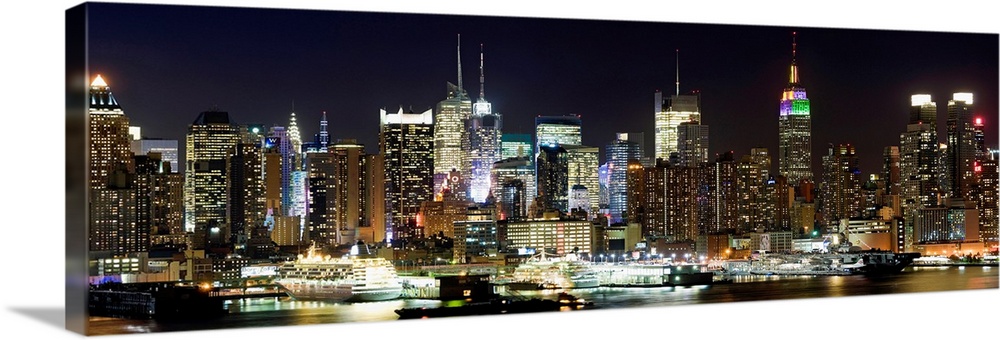 Oversized panoramic photograph of brightly lit skyscrapers in Manhattan, behind the reflecting waters of the Hudson River.