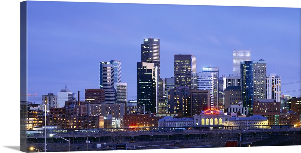 A large panoramic piece of a photograph of the Denver skyline with the buildings lit up during sun down.