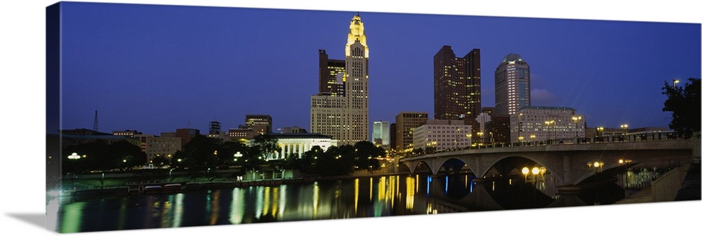 The city of Columbus Ohio is illuminated under a night sky and photographed in panoramic view from across a body of water.