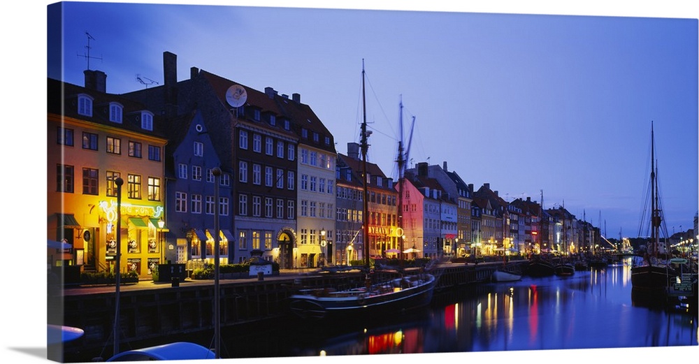 Big canvas photo of buildings along a street following a waterfront with boats lit up at night.