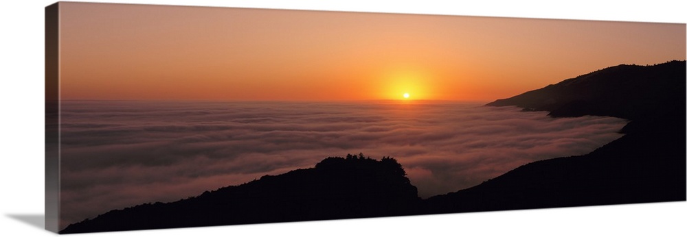A large panoramic piece of a sunset with a fog mist over the water and mountains lining the right side of the picture.