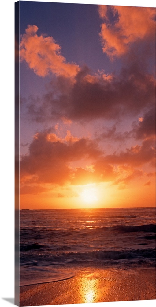 This vertical photograph shows the sun setting at the beach as waves wash against the shore.