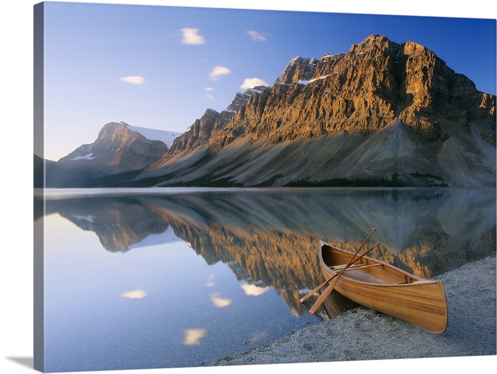 Oversized photography artwork of a canoe sitting on the edge of water with mountains in the background that reflect in the...