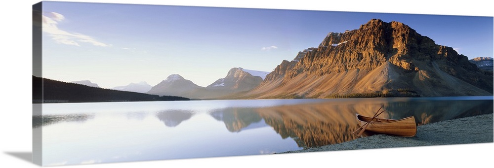 Panoramic photo on canvas of a smooth lake with a wooden canoe on the shoreline and big rocky mountains in the distance.