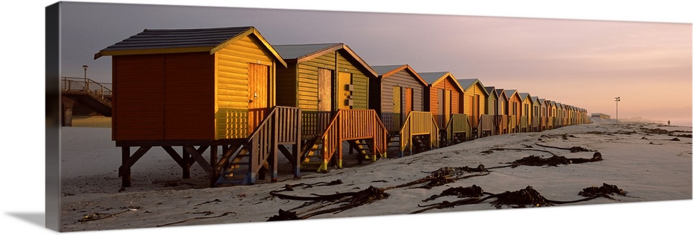 A panoramic photograph of a row of buildings built on the sandy shoreline glowing in the light of sunset.
