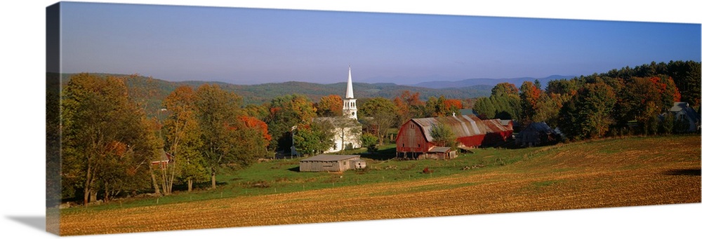 This panoramic photograph is of a barn and a church surrounded by autumn trees with large hills in the distance.