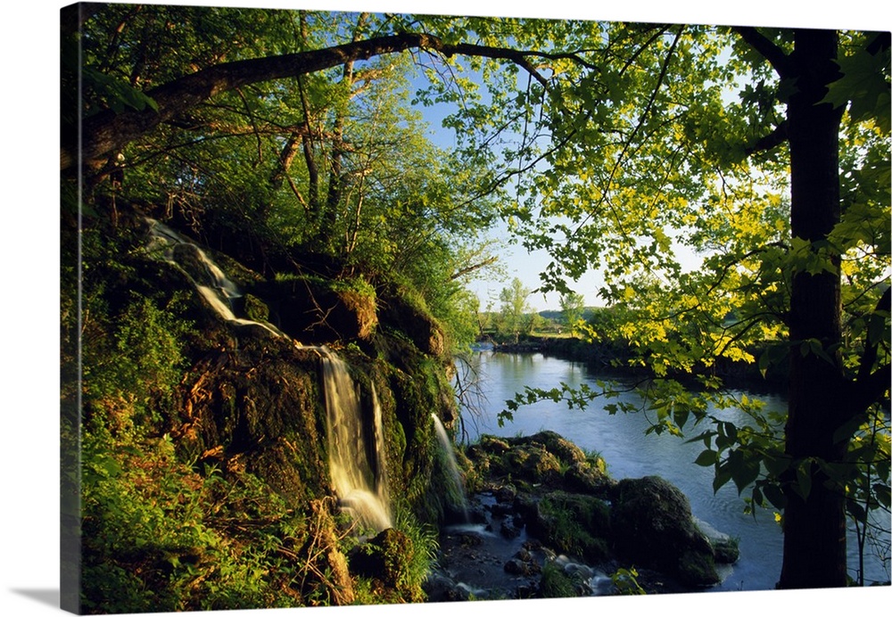 Small waterfall with mossy rocks and leafy trees on the edge of a river in the early morning.