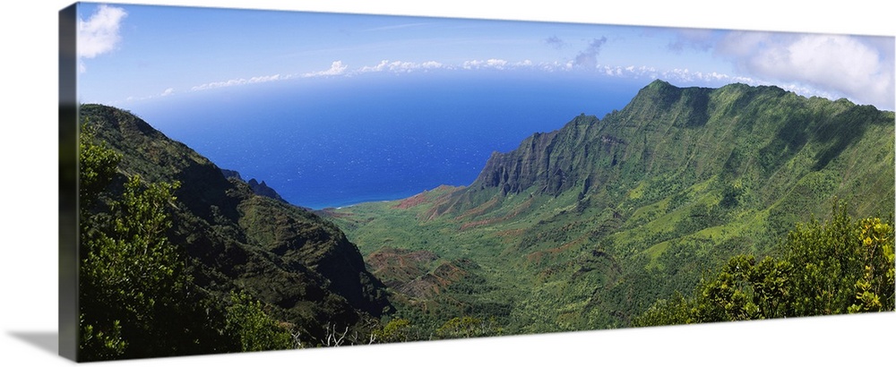 Horizontal photograph on a large wall hanging of a green cliff that overlooks the blue sea, beneath a sky with few small c...