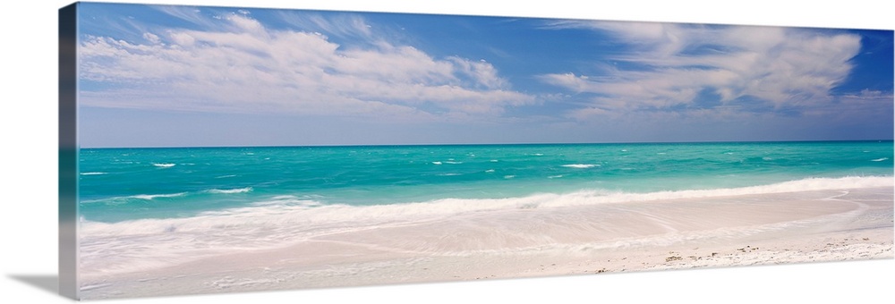 Panoramic photograph of calm ocean with surf and sand in the foreground and cloudy sky above.
