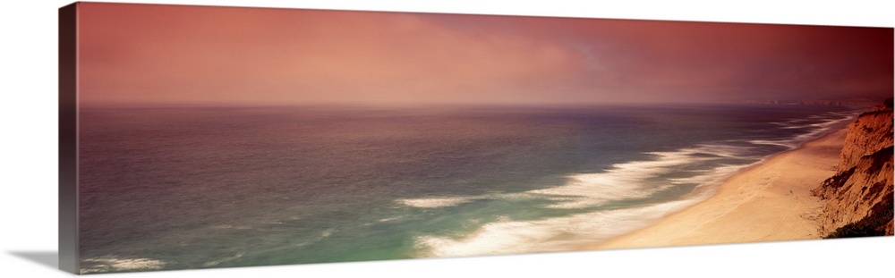 Long photo on canvas of cliffs on the right reaching a beach and crashing ocean waves on the left.