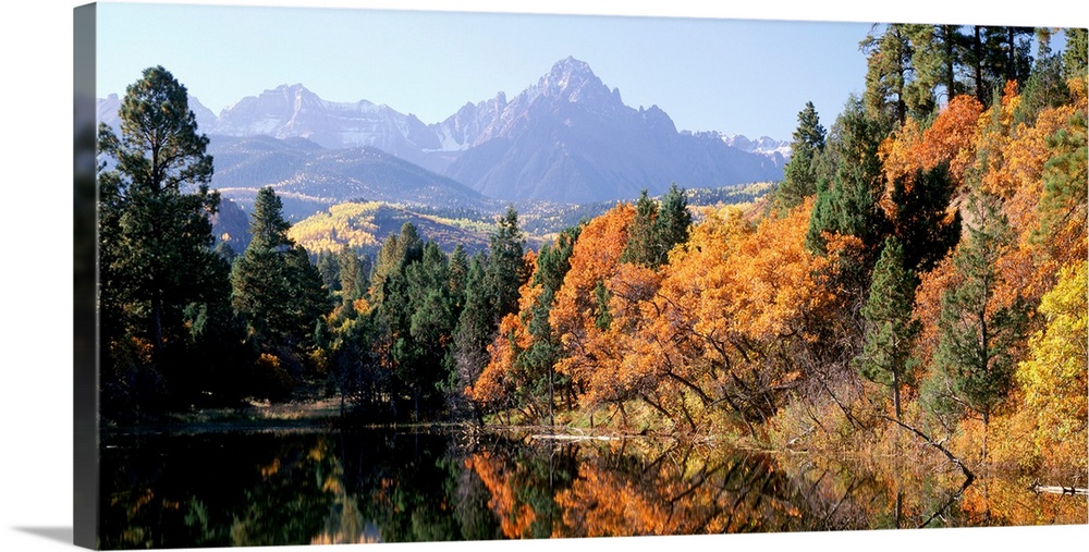 Panoramic photograph displays a colorful forest surrounding a quiet lake in Fall.  In the background there are trees leadi...