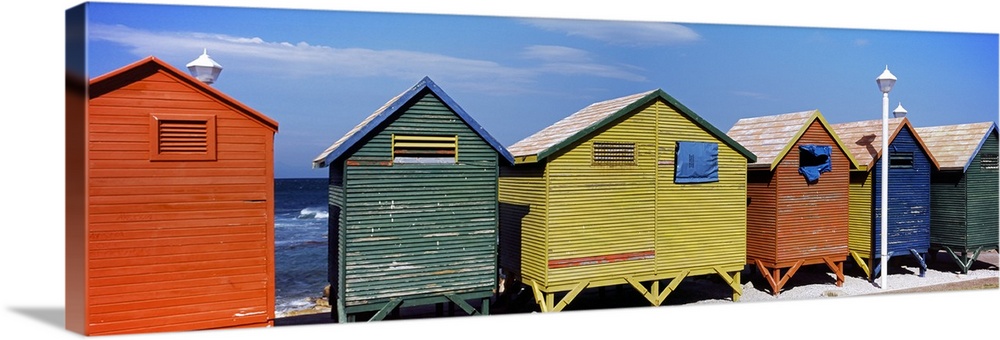 Panoramic photograph taken of multi-colored huts that line the beach with the ocean viewed in the distance.