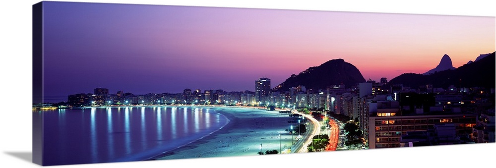 Panoramic, giant photograph of buildings lit along Copacabana Beach at night, mountains in the background against a vivid ...