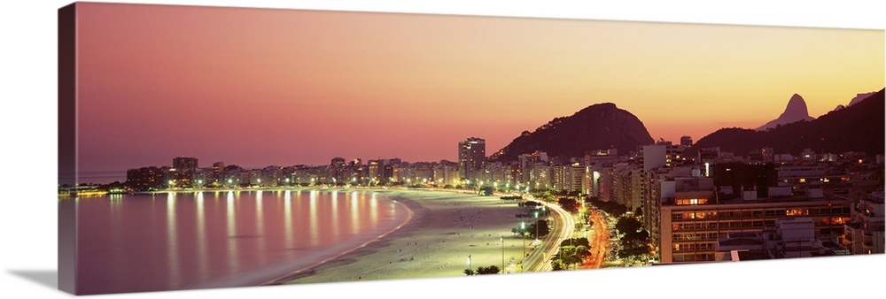 Giant landscape photograph of the curving shoreline of Copacabana Beach, lined with buildings that are lit at night, in Ri...