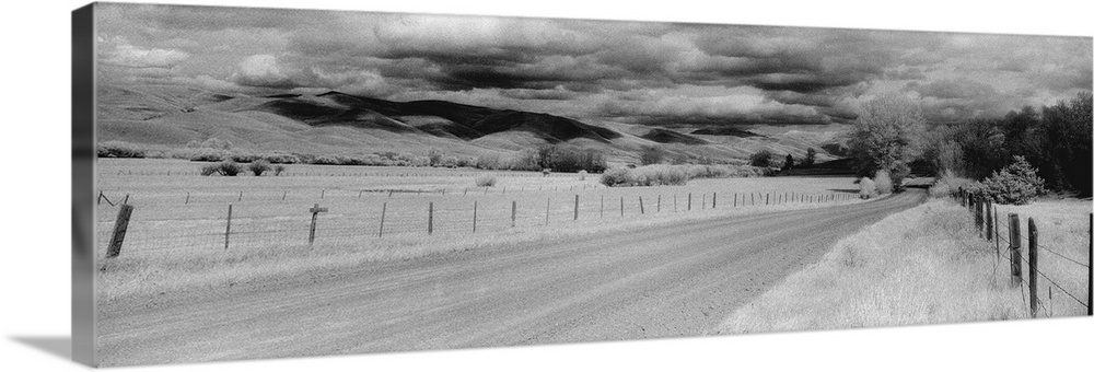 This black and white panoramic photograph is of a long dirt road that is lined on either side by a small wire fence. Large...