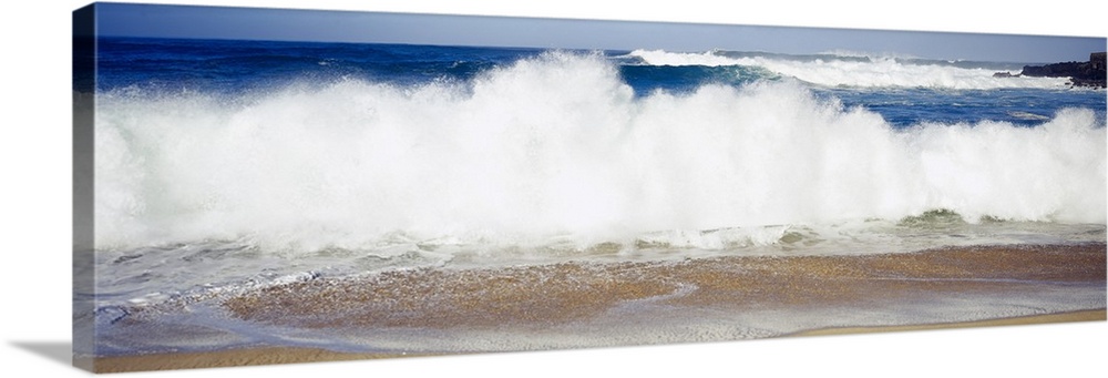 Panoramic photograph of large waves crashing into the beach, bright blue water in the background, in Waimea Bay, Hawaii.