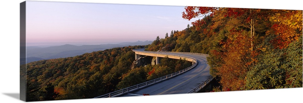 Panoramic photograph of winding mountain road with tree tops below it and mountain silhouette in the distance.