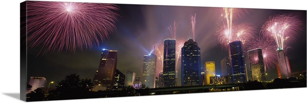 Giant photograph at nighttime displays vibrant pyrotechnics bursting above a set of large skyscrapers located within the l...