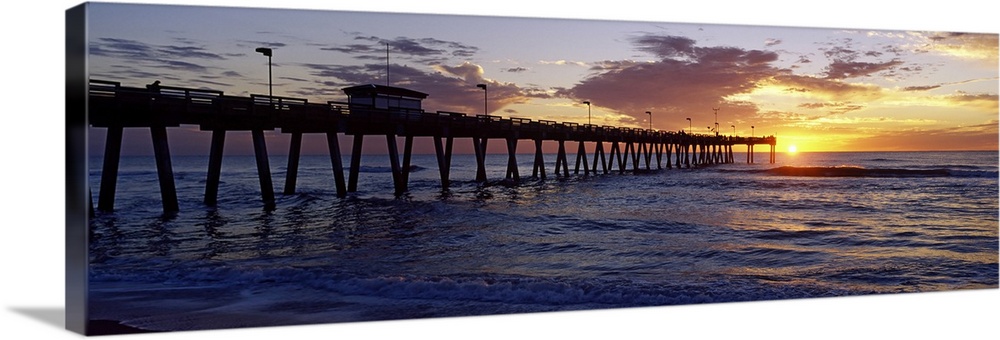 A pier that reaches far out into the ocean is photographed from the beach with the sun just setting on the horizon.
