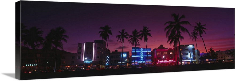 A panoramic canvas of neon lights on art deco buildings on the shore front lined with palm trees.