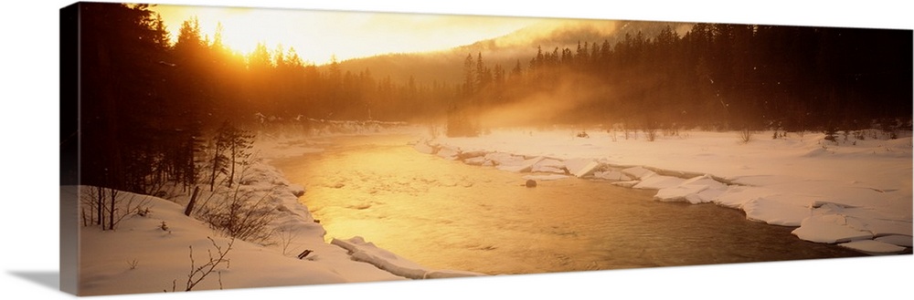 Panoramic image of a river running through a frozen landscape with fog coming off it and warm sunlight breaking through th...