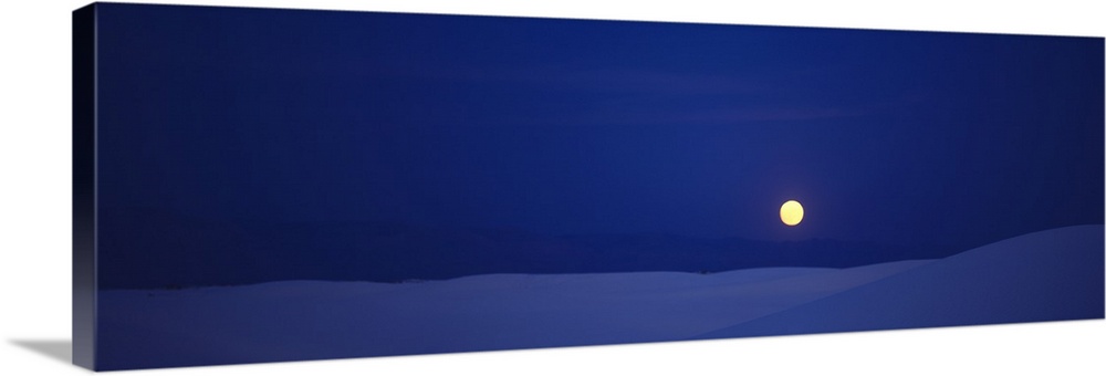 Panoramic photograph taken of a full moon against a deep blue sky and sitting above white sand that is shaded blue.