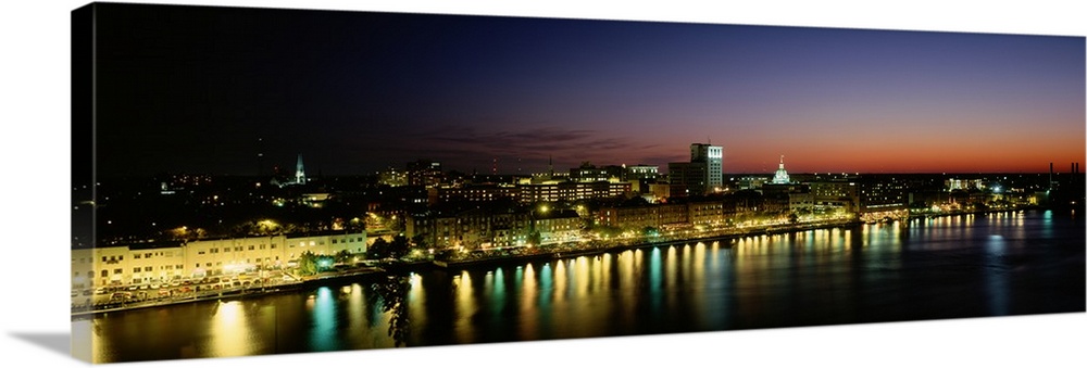 Panoramic photograph of lit up skyline and waterfront at sunset under a colorful sky.