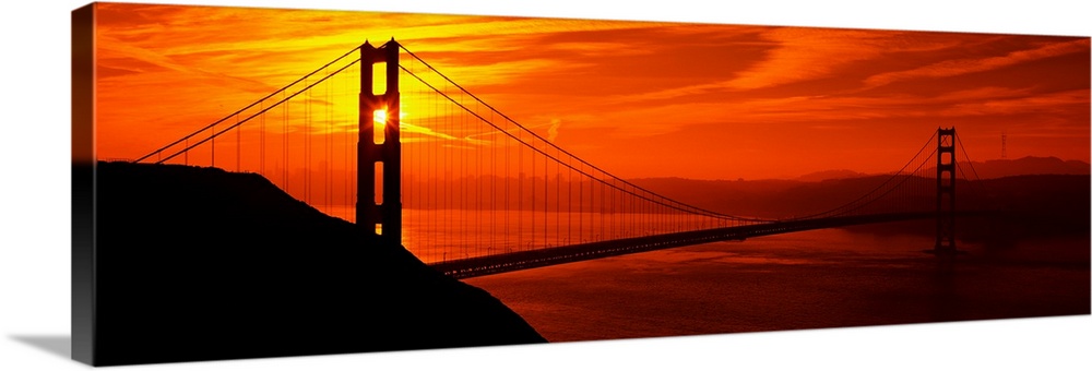 This oversized wall art is a panorama of San Francisco Bay at sunset where the suspension bridge is a dark silhouette in t...